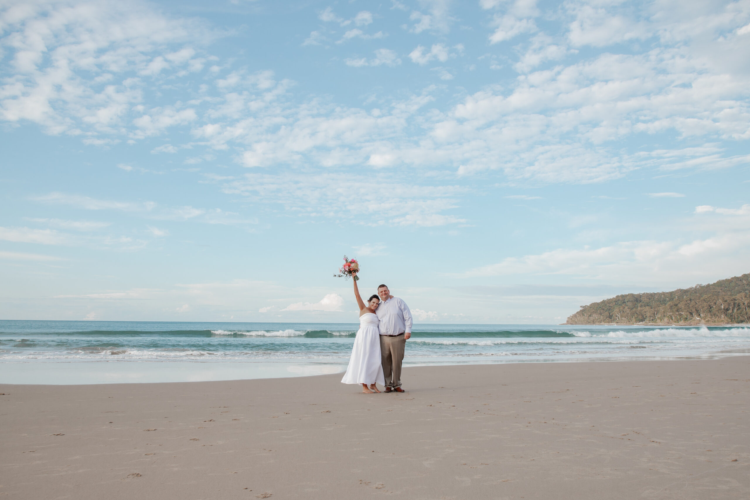 Couple on a beach dressed in wedding attire holding a bouquet of spring flowers